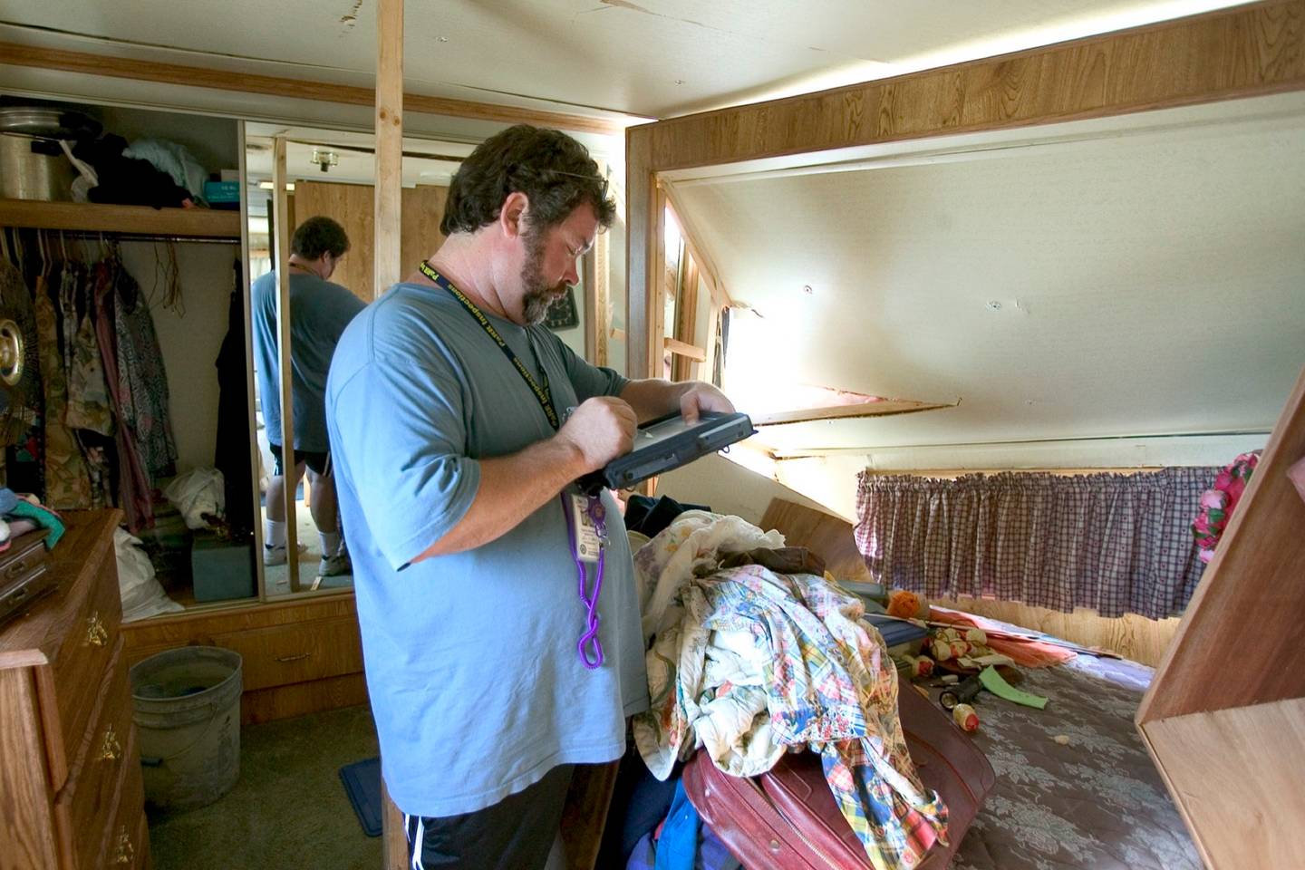 An inspector stands in a storm-damaged bedroom with a torn ceiling, recording the damage on a tablet.