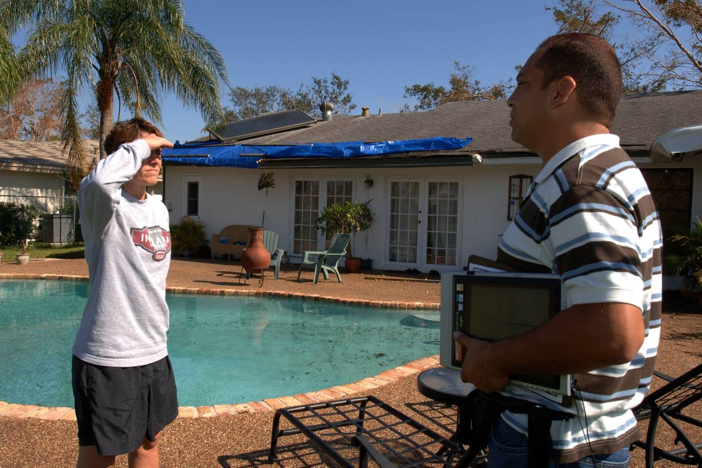 A homeowner talks with an inspector holding a tablet beside a pool, in front of a house with a blue tarp across its roof.