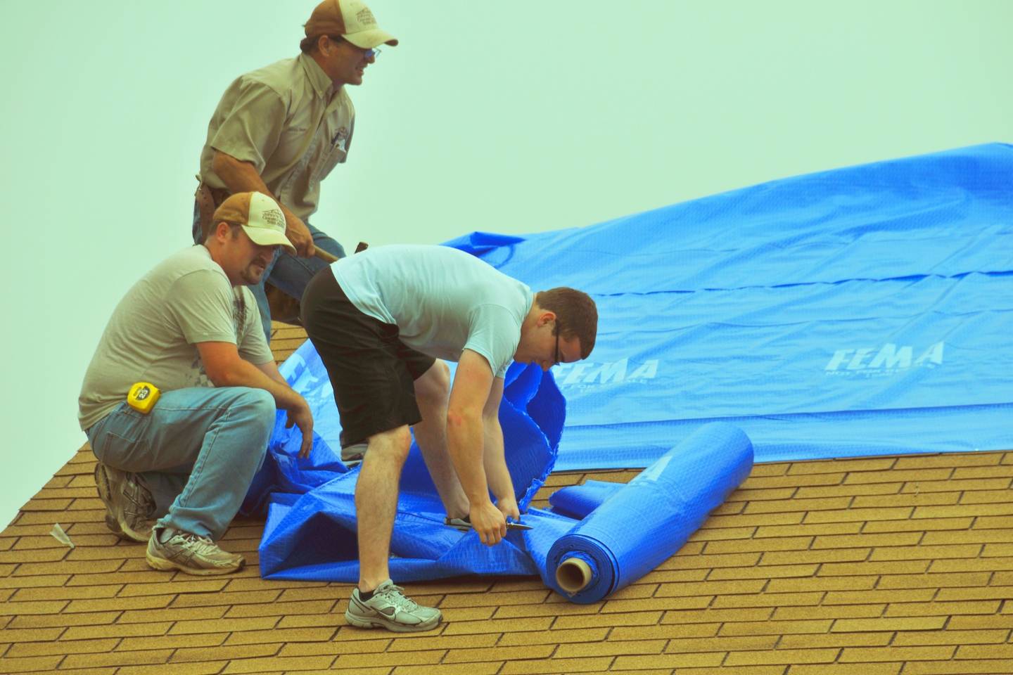 Three men on a shingled roof roll out and cut a large blue tarp to cover storm damage.