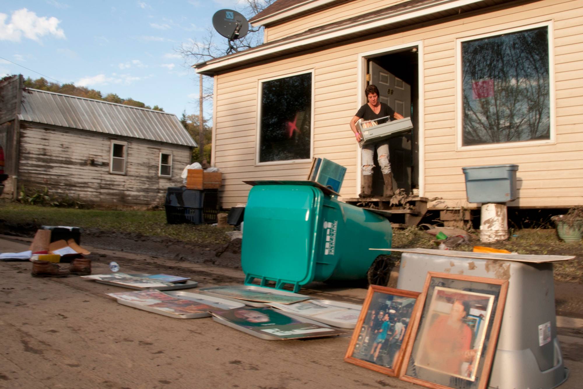 A woman carries a box out of her flood-damaged house while framed family photos and papers dry on the street in front of it.